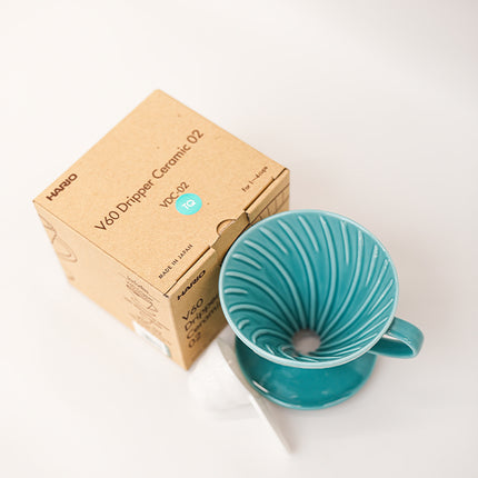 A turquoise ceramic pour-over dripper with a matching saucer and a kraft paper box labeled HARIO V60 Dripper Ceramic 02, photographed on a clean white background.